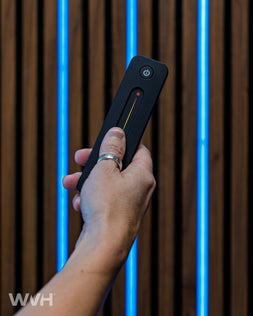 Hand holding a black remote control with a power button against a wooden slat wall background with blue LED light strips